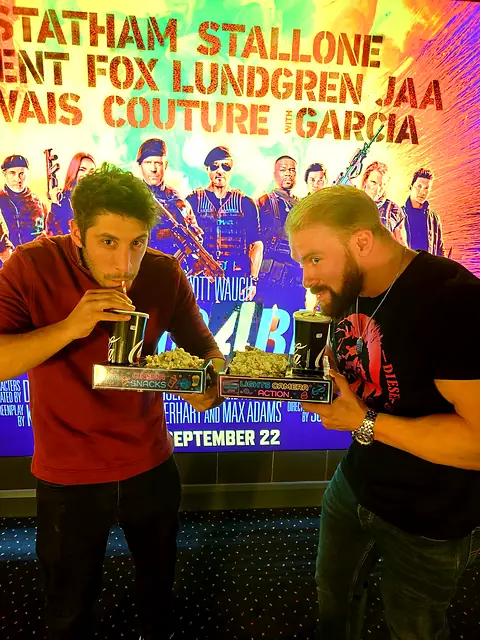 Two men holding neon snack trays in front of a cinema film poster