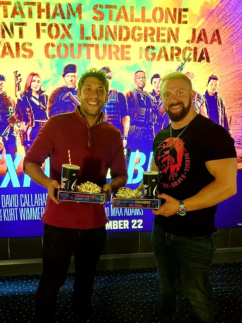 Two men with neon snack trays and drinks beside a cinema film poster